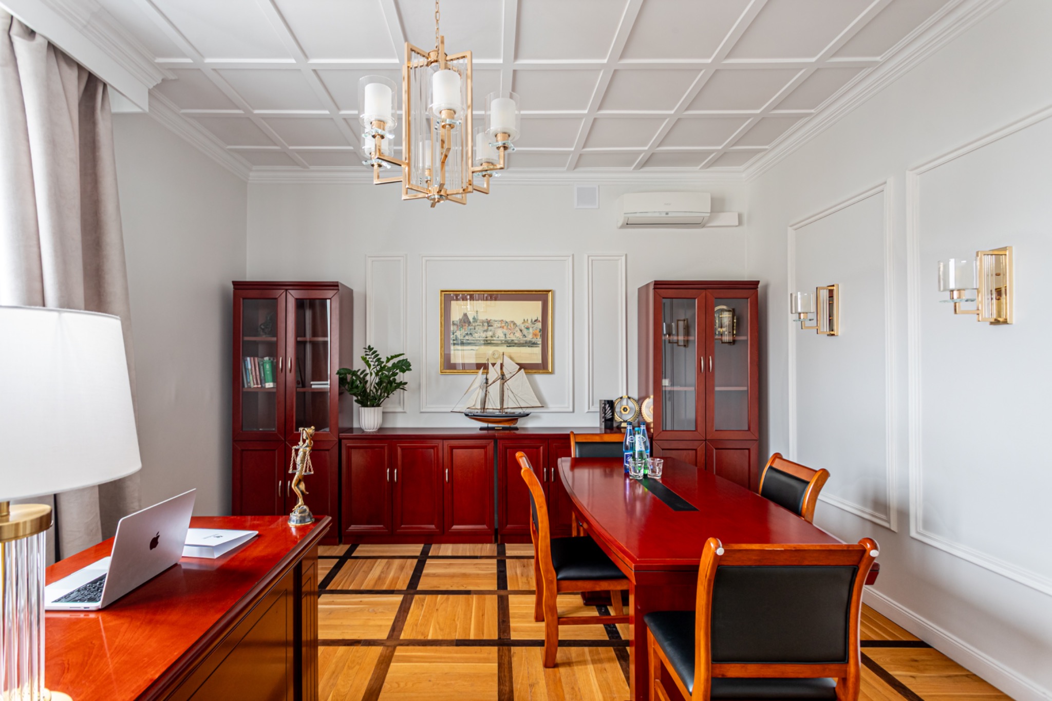 Eleganckie wnętrze gabinetu z mahoniowymi meblami, parkietem w szachownicę i dekoracyjnym oświetleniem. Na biurku laptop i statuetka Temidy.