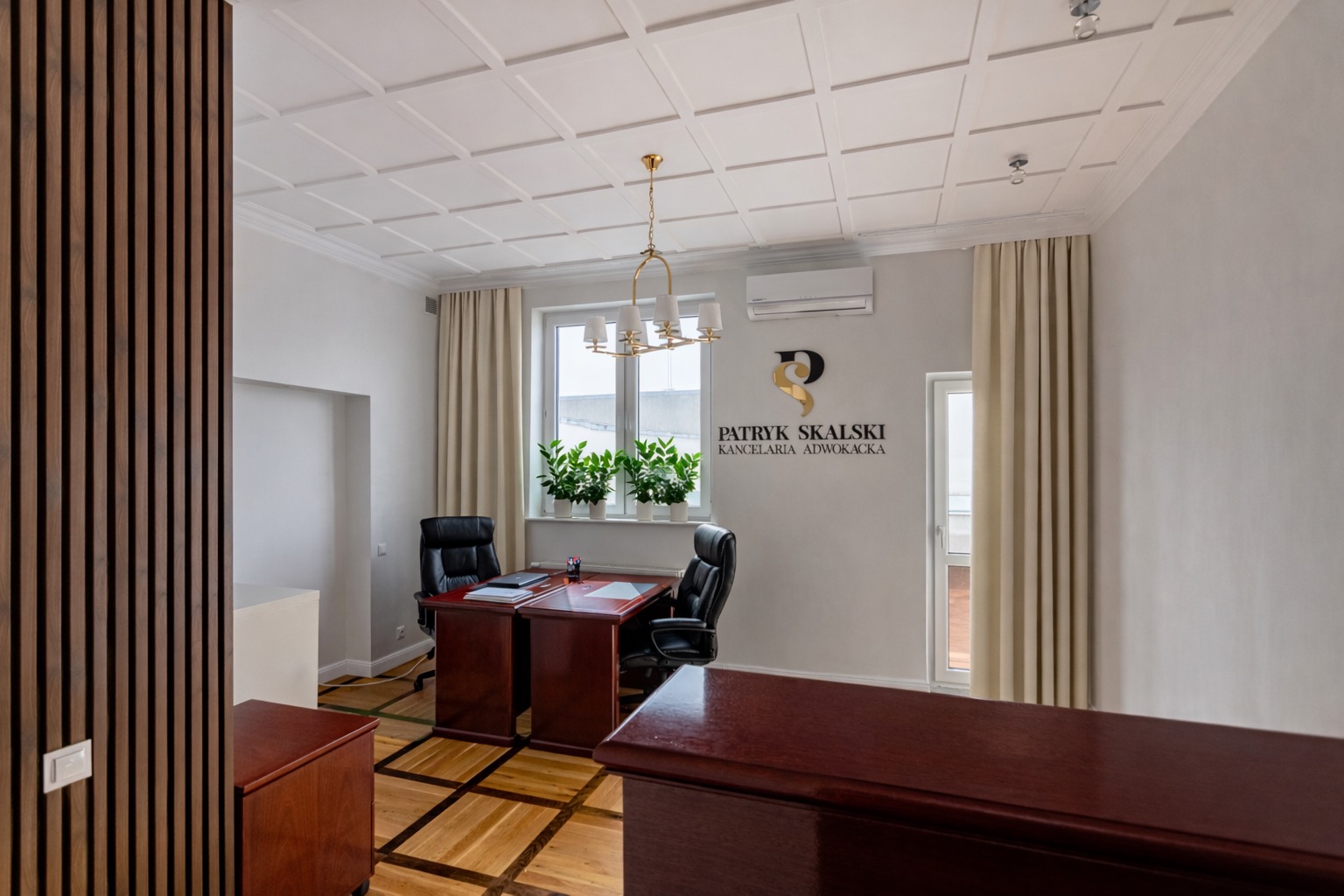 Eleganckie wnętrze kancelarii adwokackiej z drewnianymi meblami, parkietem i logo na ścianie. Widok na biurko, krzesła i okno z roślinami. Oświetlenie sufitowe.