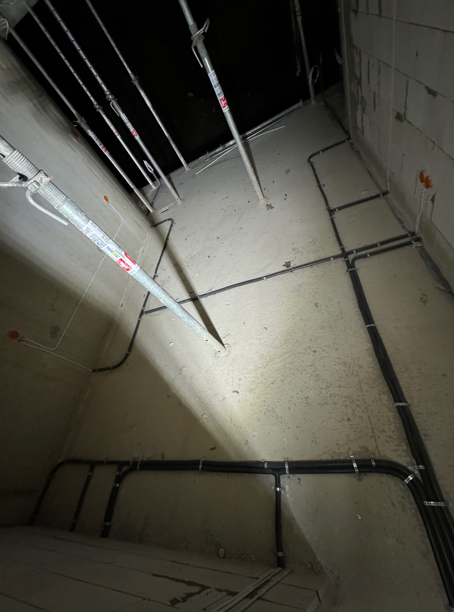 Surowy beton, wsporniki stropowe i czarne kable elektryczne ułożone wzdłuż ścian w trakcie instalacji. Widoczne pomarańczowe puszki elektryczne.