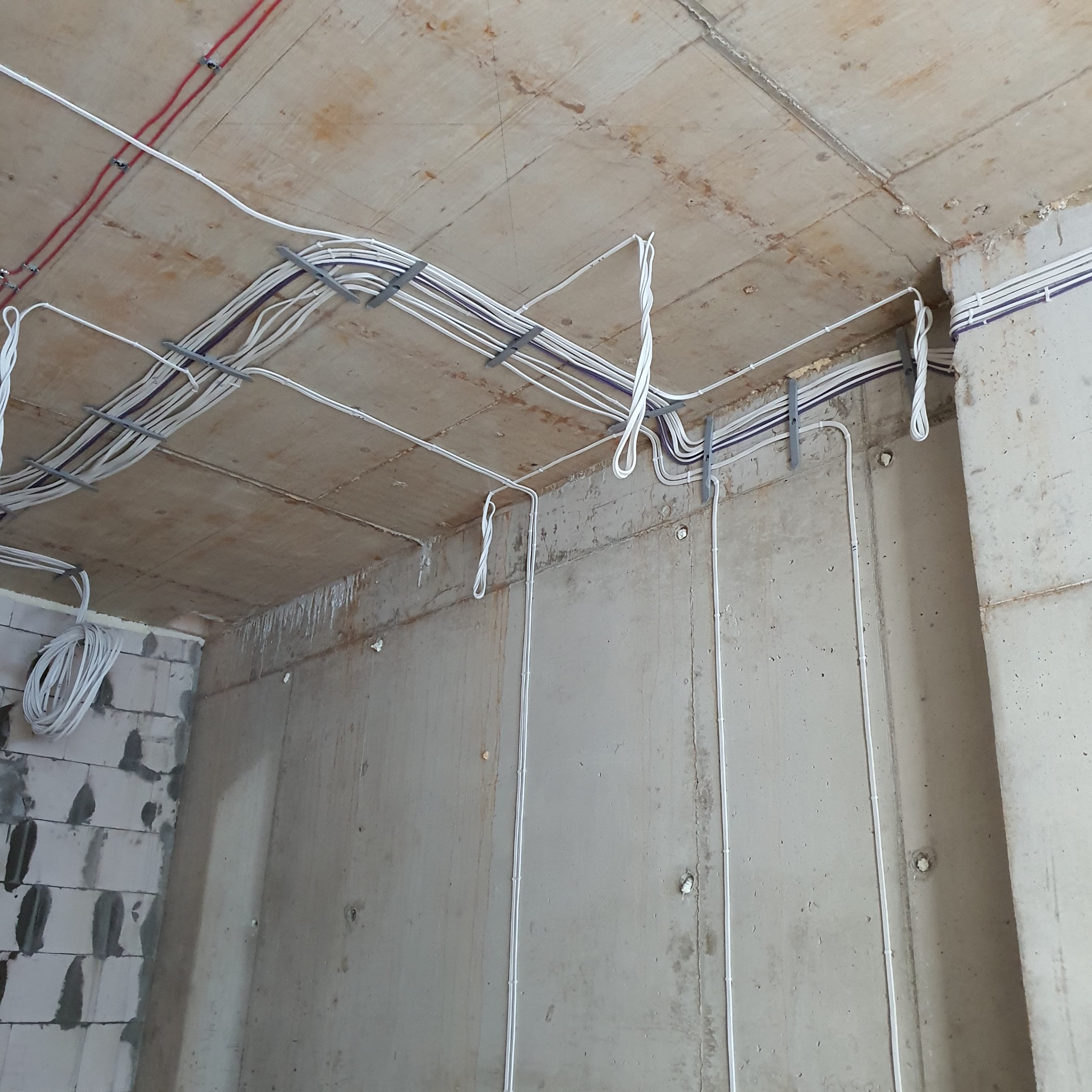 Surowy strop z betonową fakturą i fragmentem ściany, na których zamontowano kable elektryczne w peszlach, przymocowane metalowymi uchwytami.