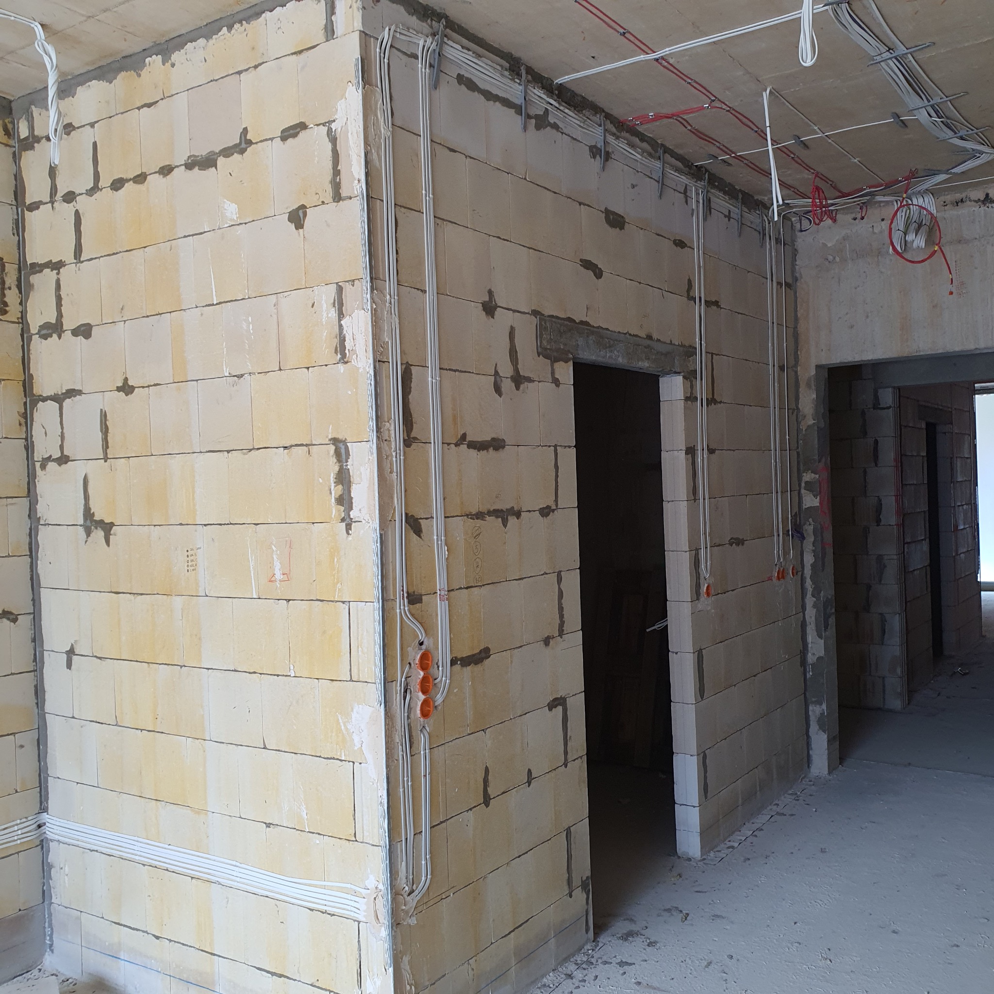 Instalacja elektryczna w surowym wnętrzu: białe przewody w peszlach prowadzone po ścianach z bloczków, widoczne puszki instalacyjne i kable pod sufitem.