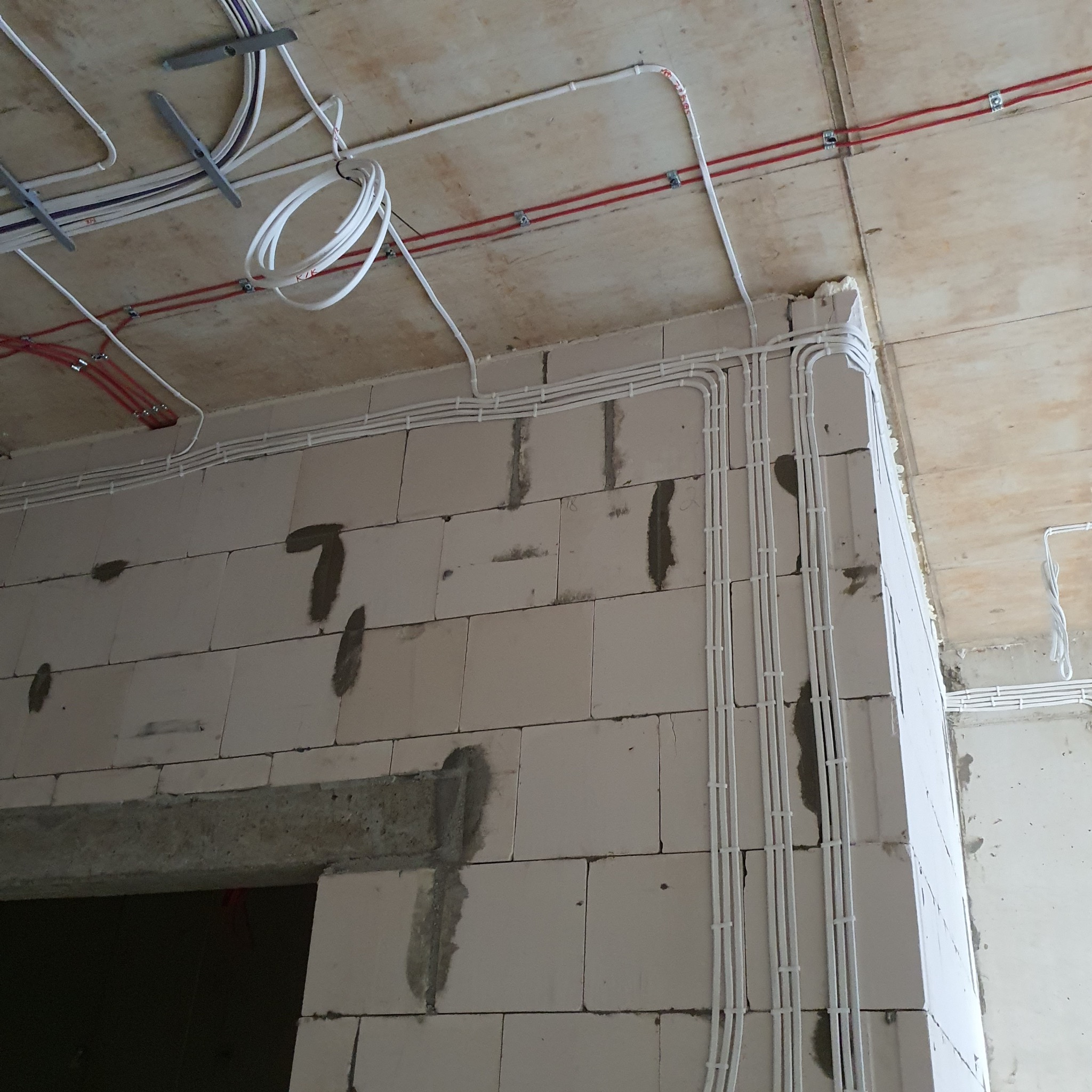 Białe kable elektryczne równolegle ułożone na ścianie z betonu komórkowego, widoczne mocowania i fragmenty instalacji pod stropem. Surowy stan wykończenia.