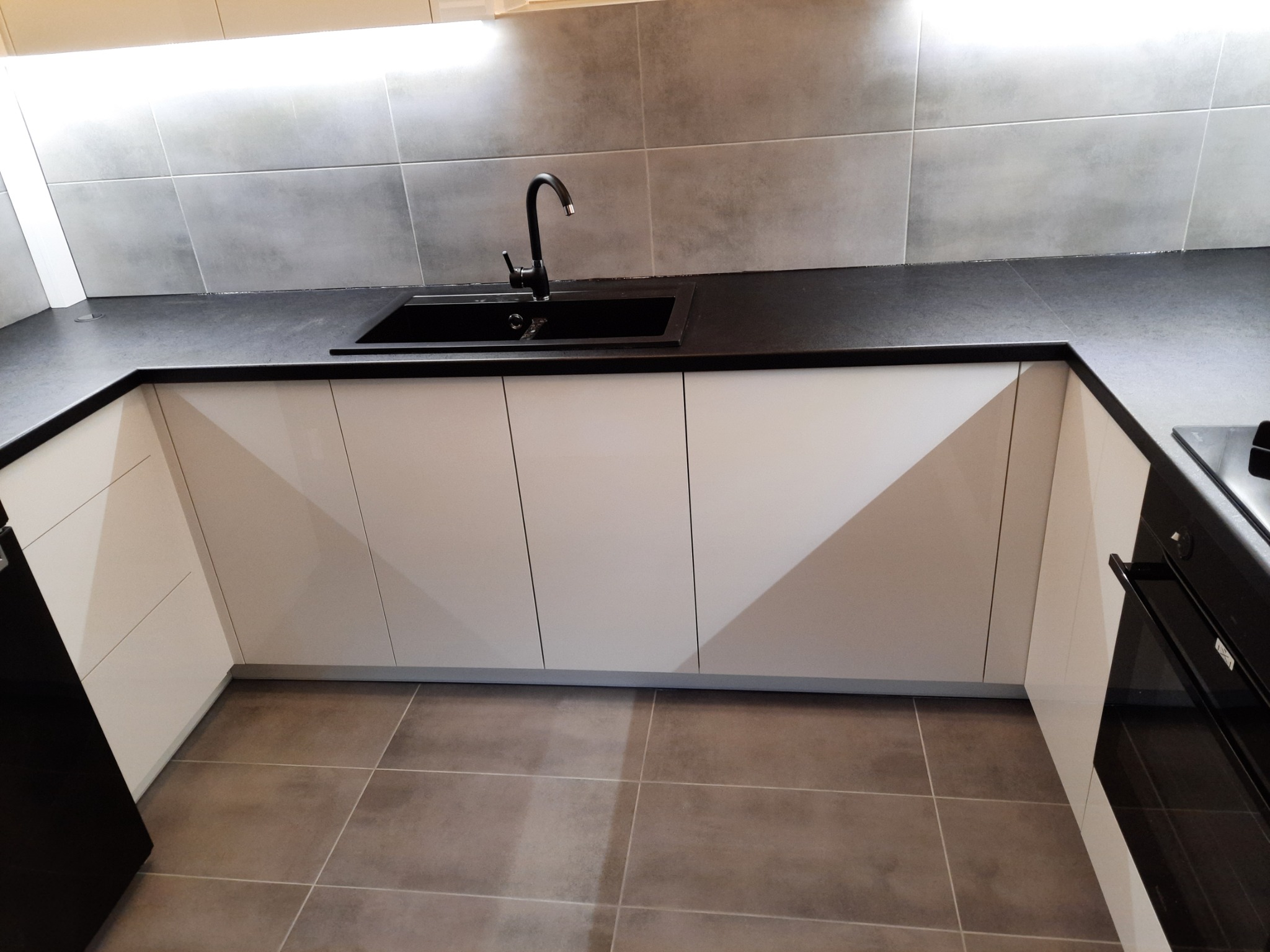 Nowoczesna kuchnia z białymi szafkami dolnymi o geometrycznym wzorze, czarnym blatem i zlewozmywakiem, szarymi płytkami na ścianie i podłodze, oświetleniem podszafkowym.