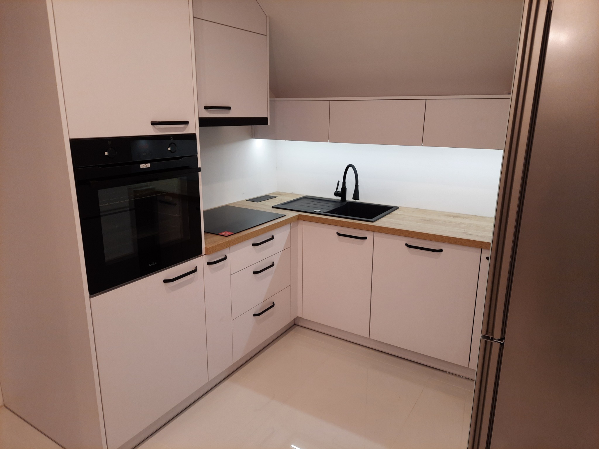 Nowoczesna kuchnia w jasnych barwach z drewnianym blatem, czarnym zlewem i minimalistycznymi uchwytami, oświetlona od dołu, z piekarnikiem marki Amica.