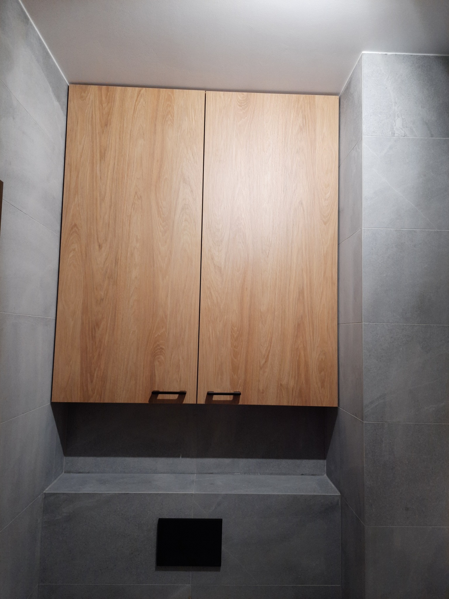 Zamknięta szafka wisząca z drewnopodobnymi frontami i ciemnymi uchwytami w łazience wyłożonej szarymi płytkami.