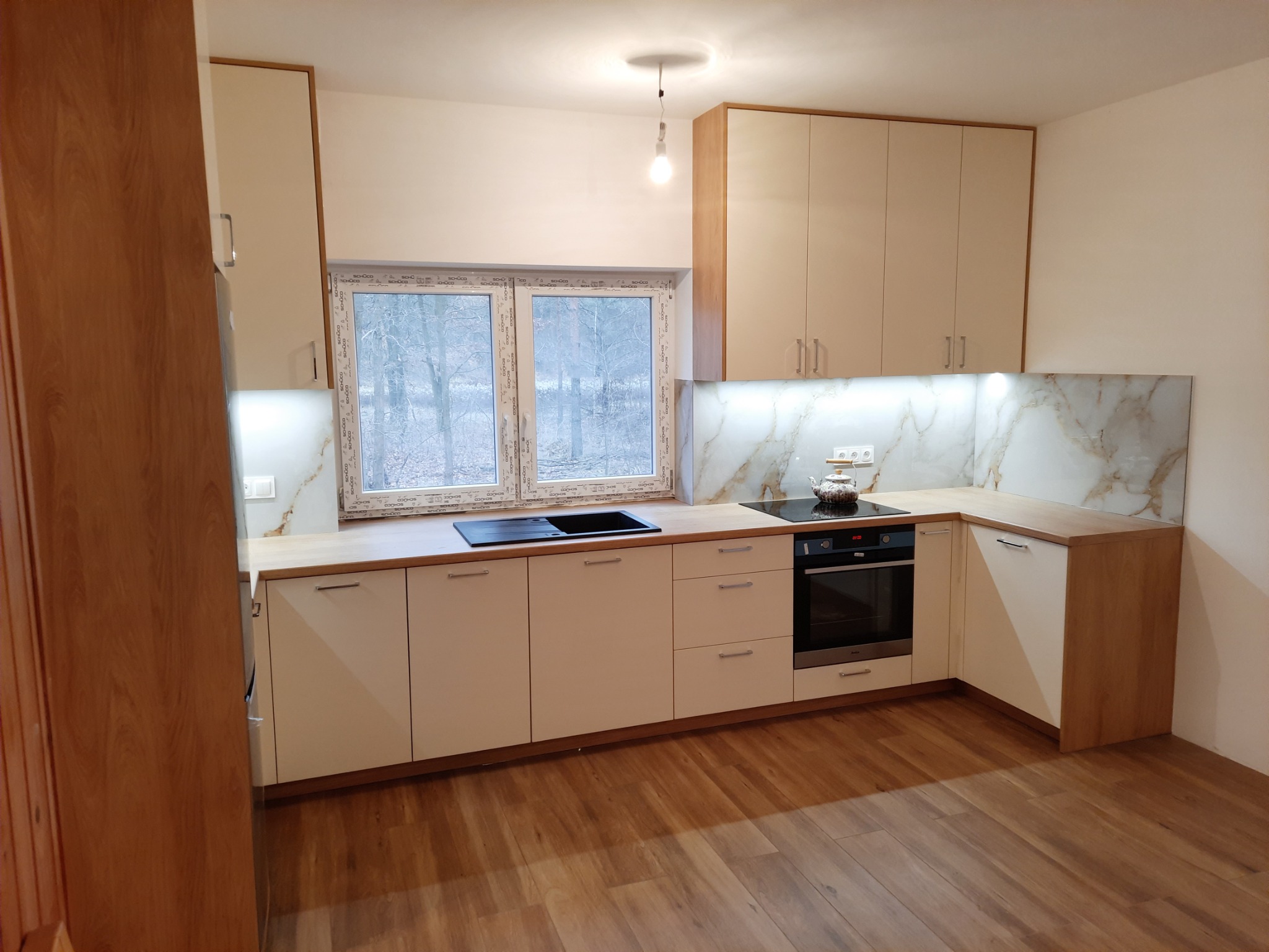Nowoczesna kuchnia z beżowymi frontami szafek, drewnianymi blatami i marmurkowym panelem nad blatem, widok na okno z zimowym krajobrazem.