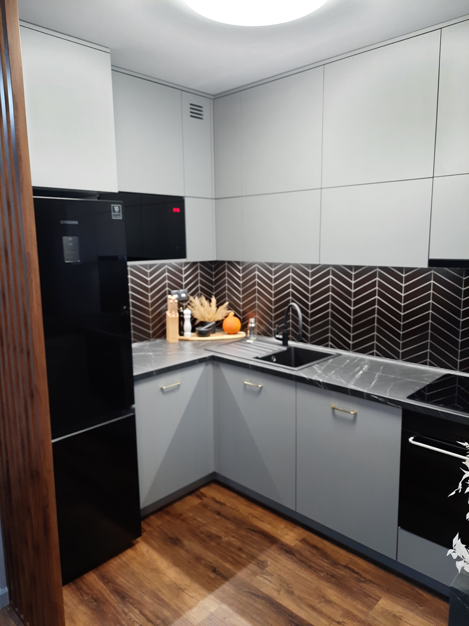 Nowoczesna kuchnia z szarymi szafkami, czarnym blatem i chevronowym backsplashiem, uzupełniona czarną lodówką i drewnianą podłogą.