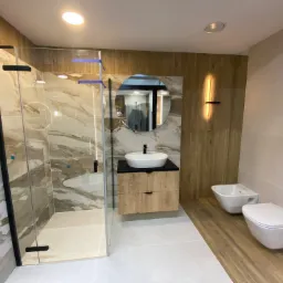 Nowoczesna łazienka z kabiną prysznicową typu walk-in z czarnymi profilami, umywalką nablatową na drewnianej szafce, okrągłym lustrem i podwieszanym bidetem oraz toaletą.