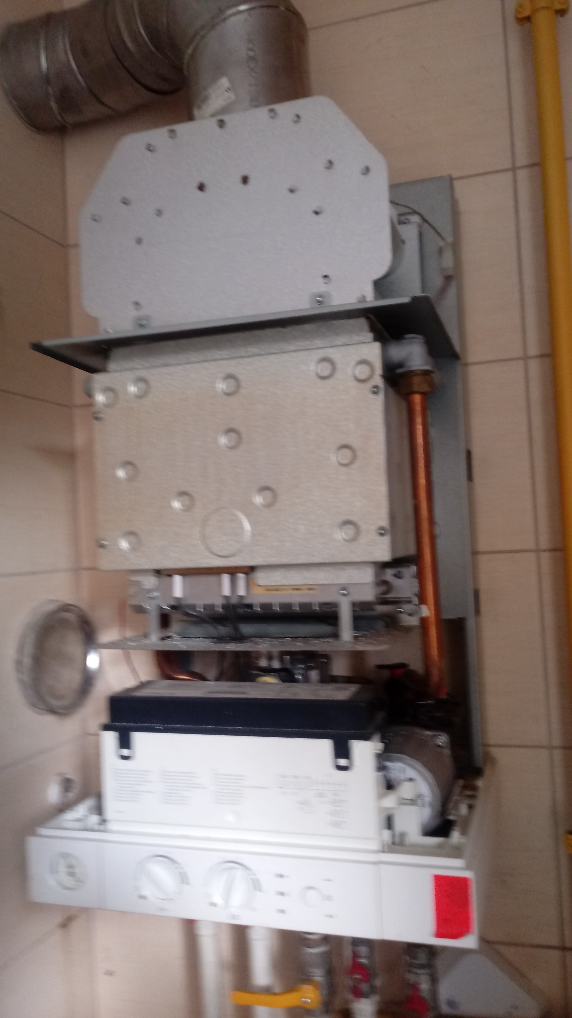 Otwarty piec gazowy wiszący na ścianie z beżowych płytek, widoczne elementy wewnętrzne, rury miedziane i odprowadzenie spalin.