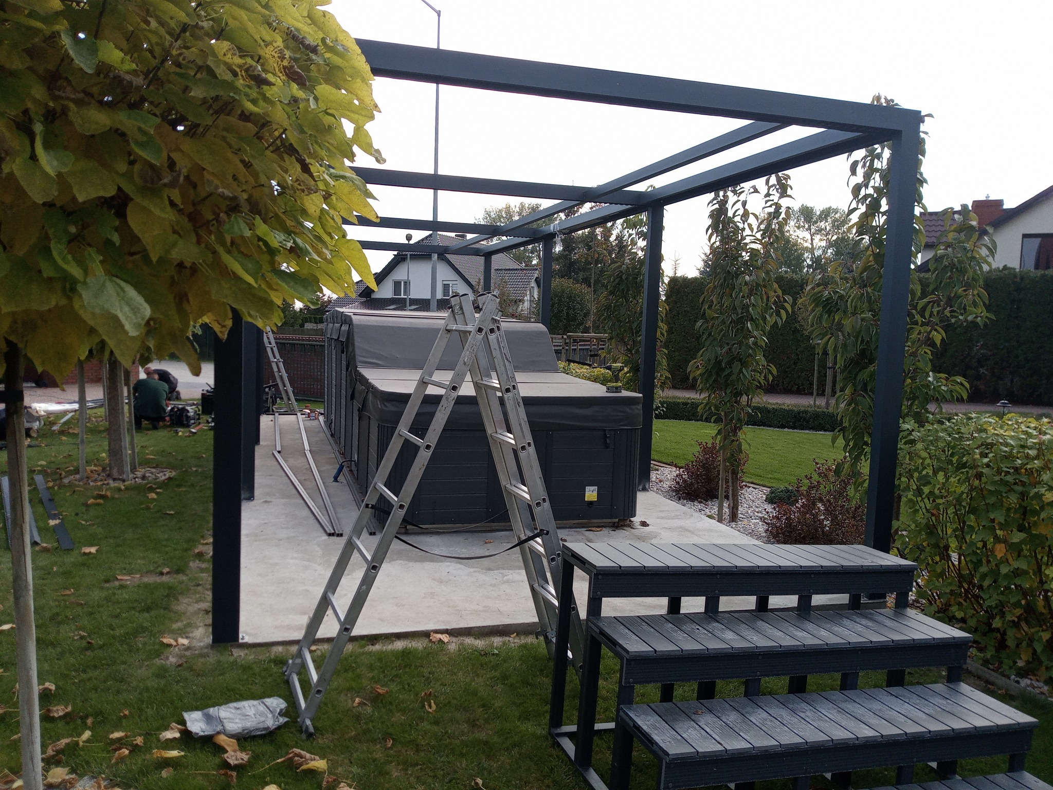 Montaż zadaszenia nad jacuzzi w ogrodzie, widoczne elementy konstrukcji, drabina i schodki z tworzywa sztucznego, trawnik z opadłymi liśćmi.