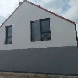 Dwukolorowa elewacja domu jednorodzinnego z ciemnoszarym dołem i jasnoszarą górą, widoczne okna z czarnymi ramami i czerwona dachówka.