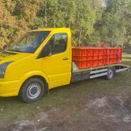 Żółty samochód dostawczy z platformą załadowaną pomarańczowymi elementami konstrukcyjnymi, zaparkowany na trawiastym terenie w jesiennej scenerii.