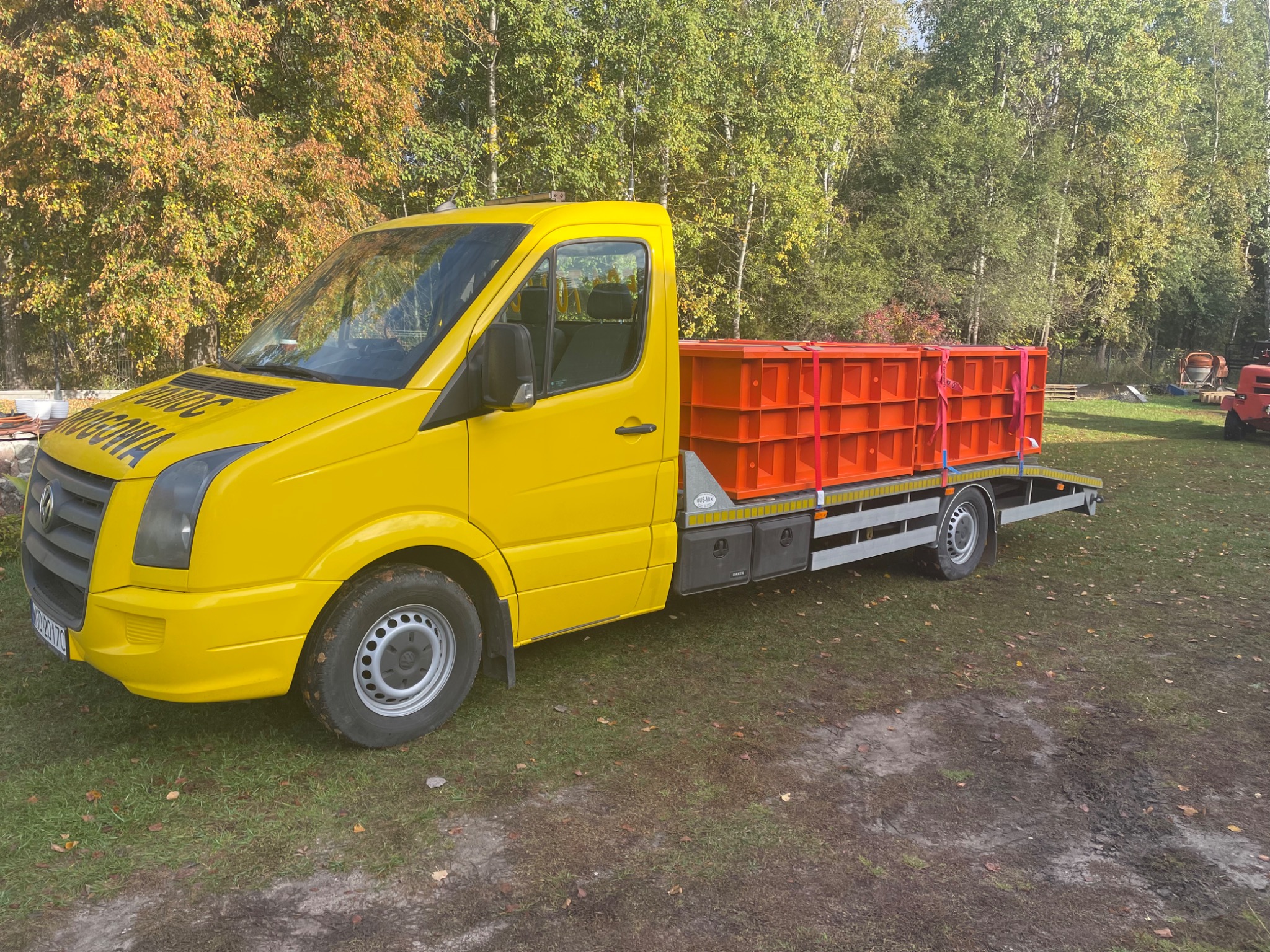 Żółty samochód dostawczy z platformą załadowaną pomarańczowymi elementami konstrukcyjnymi, zaparkowany na trawiastym terenie w jesiennej scenerii.