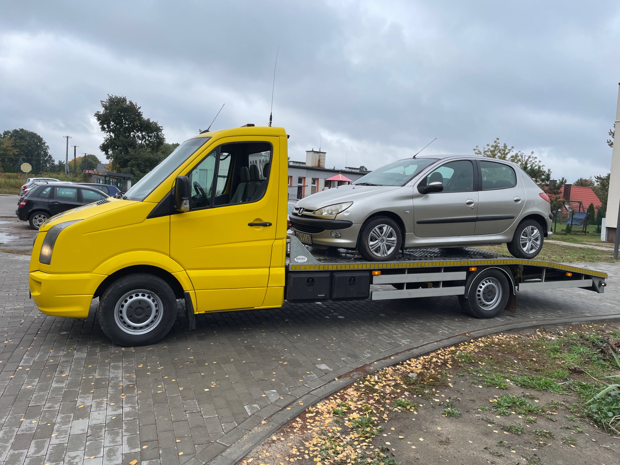 Żółta laweta z załadowanym srebrnym samochodem osobowym na chodniku z kostki brukowej, widok z boku na tle budynków i drzew pod pochmurnym niebem.