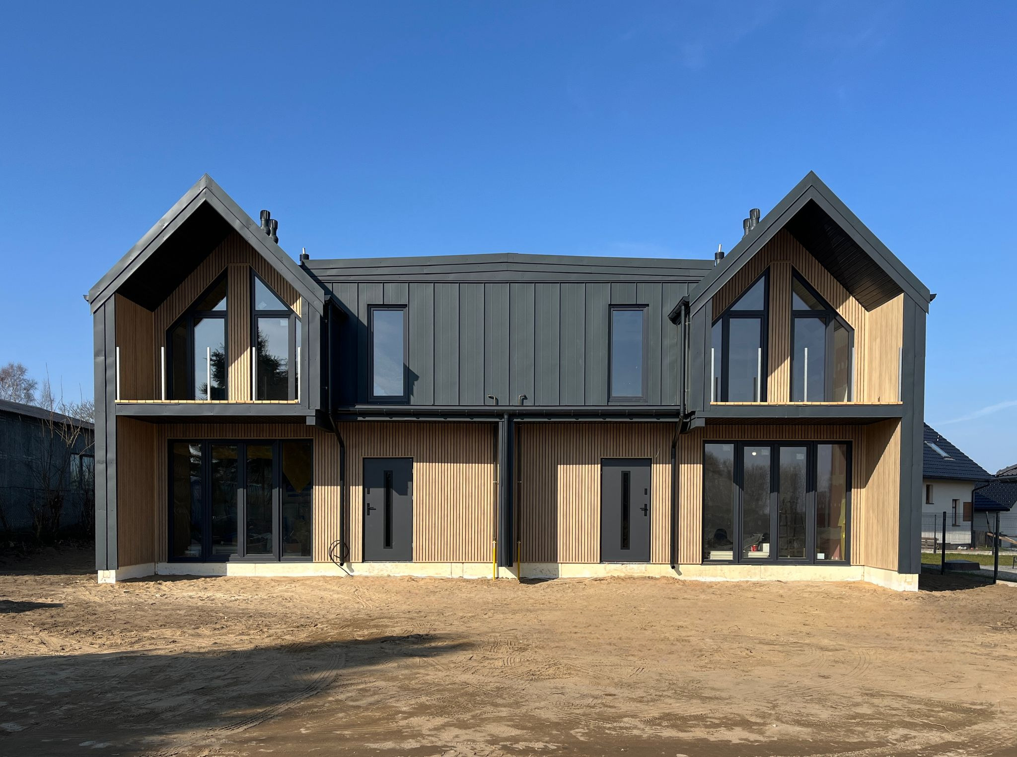 Nowoczesny bliźniak z drewnianą elewacją i grafitowymi elementami, w surowym stanie deweloperskim, na tle błękitnego nieba w słoneczny dzień.