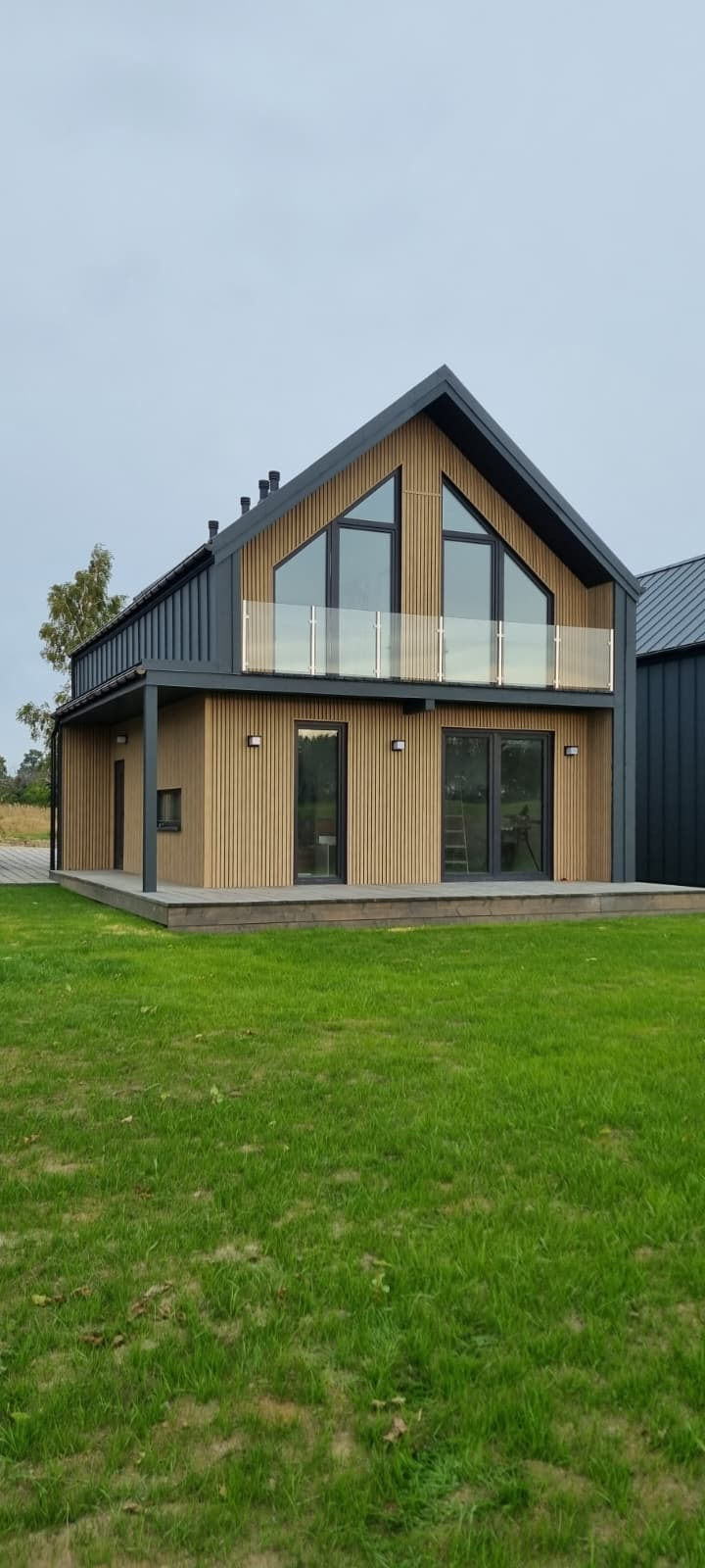 Nowoczesny dom jednorodzinny z drewnianą elewacją i balkonem z balustradą ze szkła, widok z zewnątrz na tle zielonego trawnika.