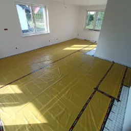 Żółta folia grzewcza rozłożona na podłodze w jasnym pokoju z dwoma oknami, przygotowana do dalszych prac wykończeniowych, widoczne taśmy montażowe z logo Termofol.