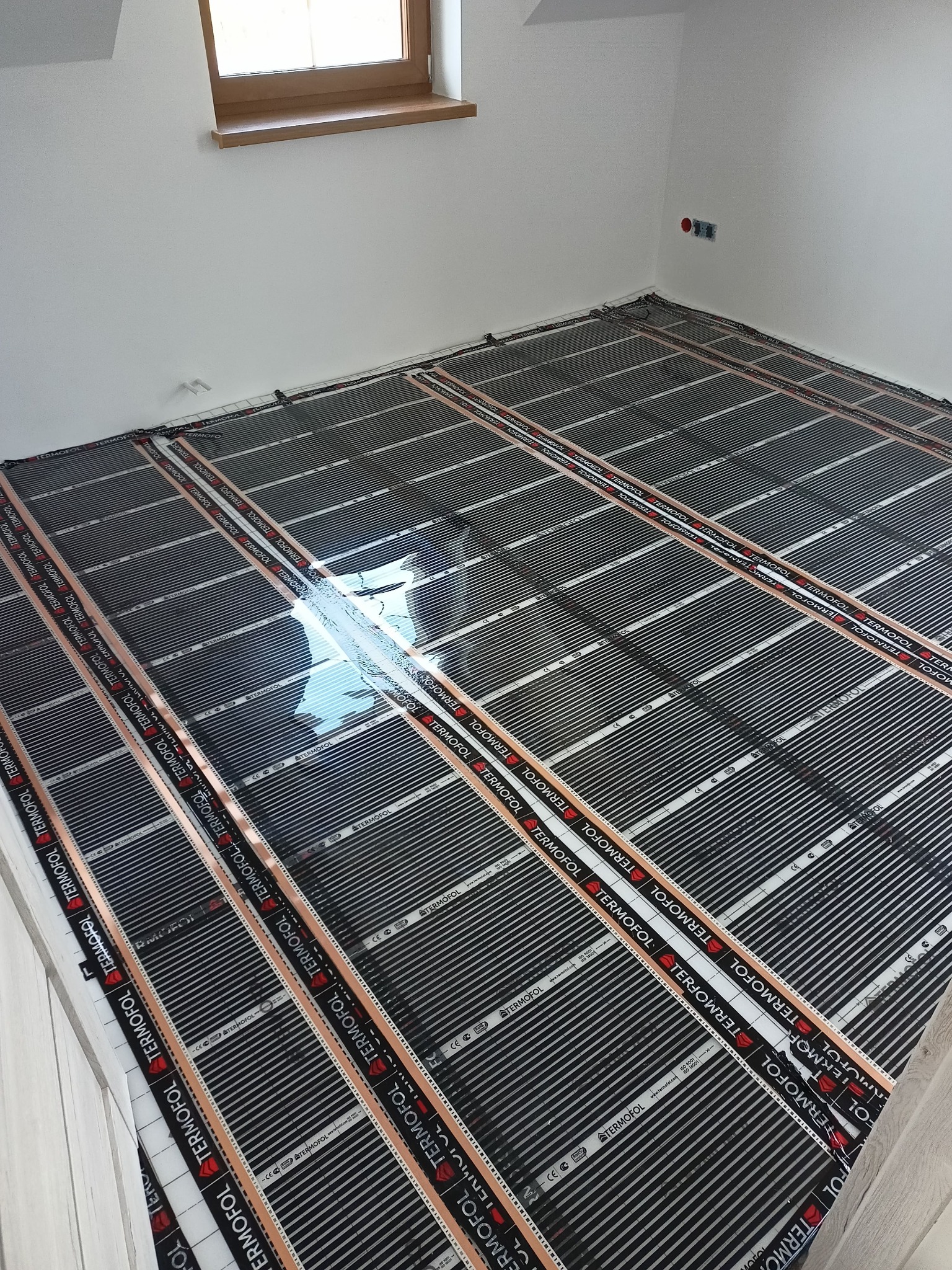 Montaż mat grzewczych Foliotermika na podłodze w jasnym pomieszczeniu z oknem, widoczne czarne maty z napisami TERMOFOL i miedzianymi paskami, częściowo przykryte folią ochronną.