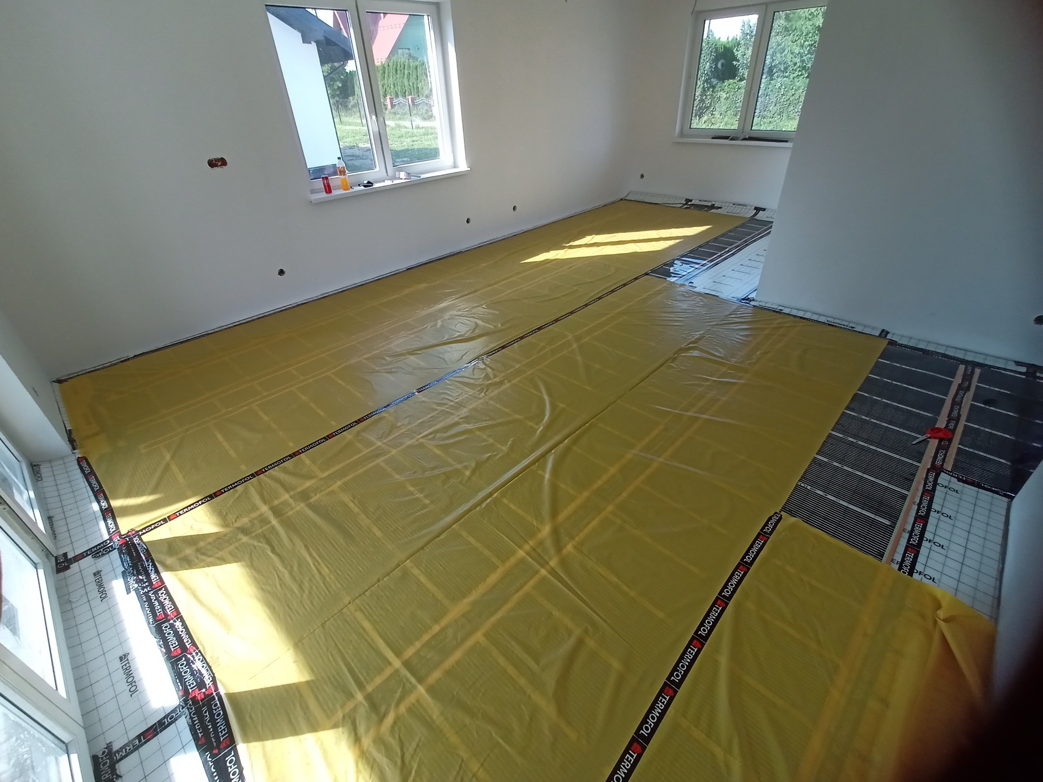 Rozłożona folia ochronna Termofol na matach grzewczych w jasnym pomieszczeniu z dwoma oknami, przygotowana do dalszych prac wykończeniowych.