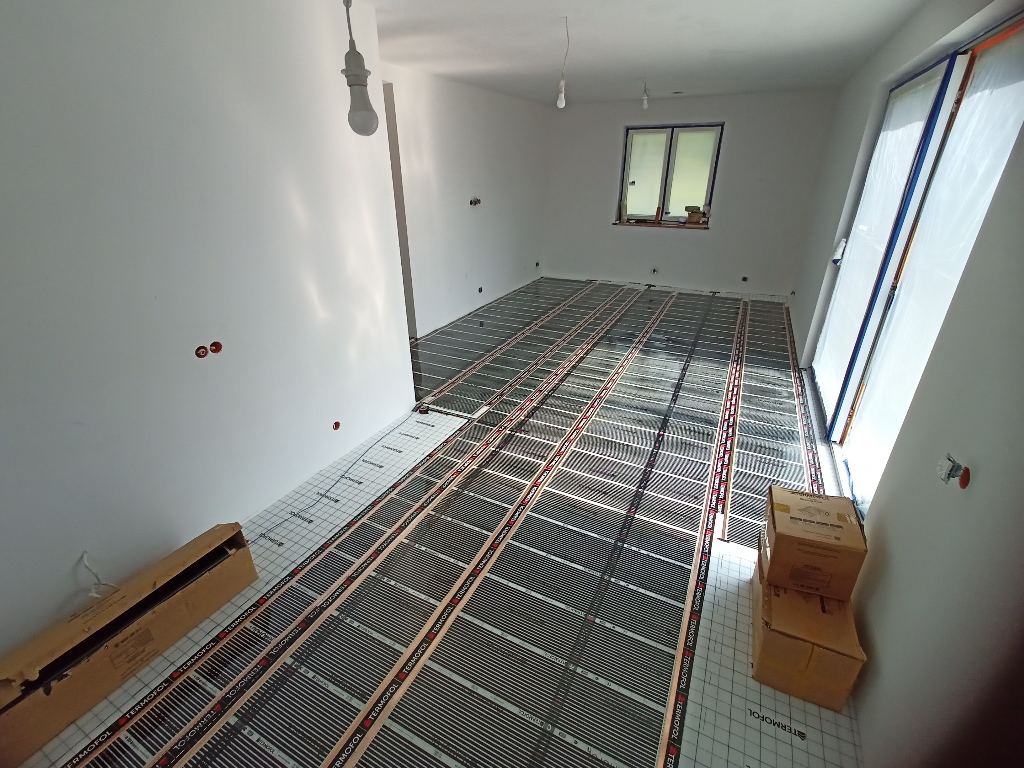 Montaż folii grzewczych marki Termofol na podłodze w nowym, jasnym pomieszczeniu z oknem zabezpieczonym folią malarską, widoczne kartony z materiałami.