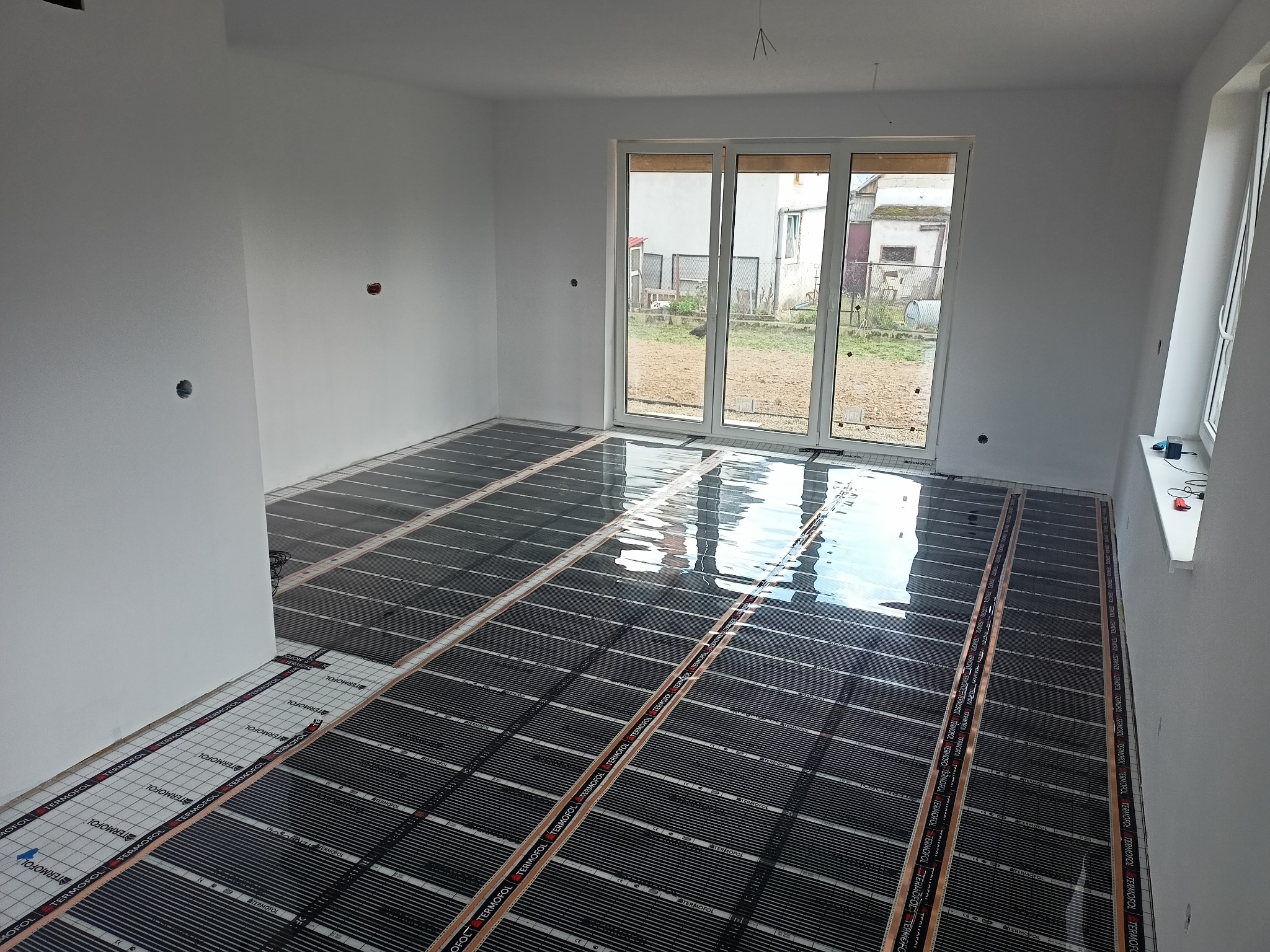 Rozłożone folie grzewcze Termofol na podłożu w nowym, jasnym pomieszczeniu z dużym oknem balkonowym, gotowe do dalszych prac wykończeniowych.