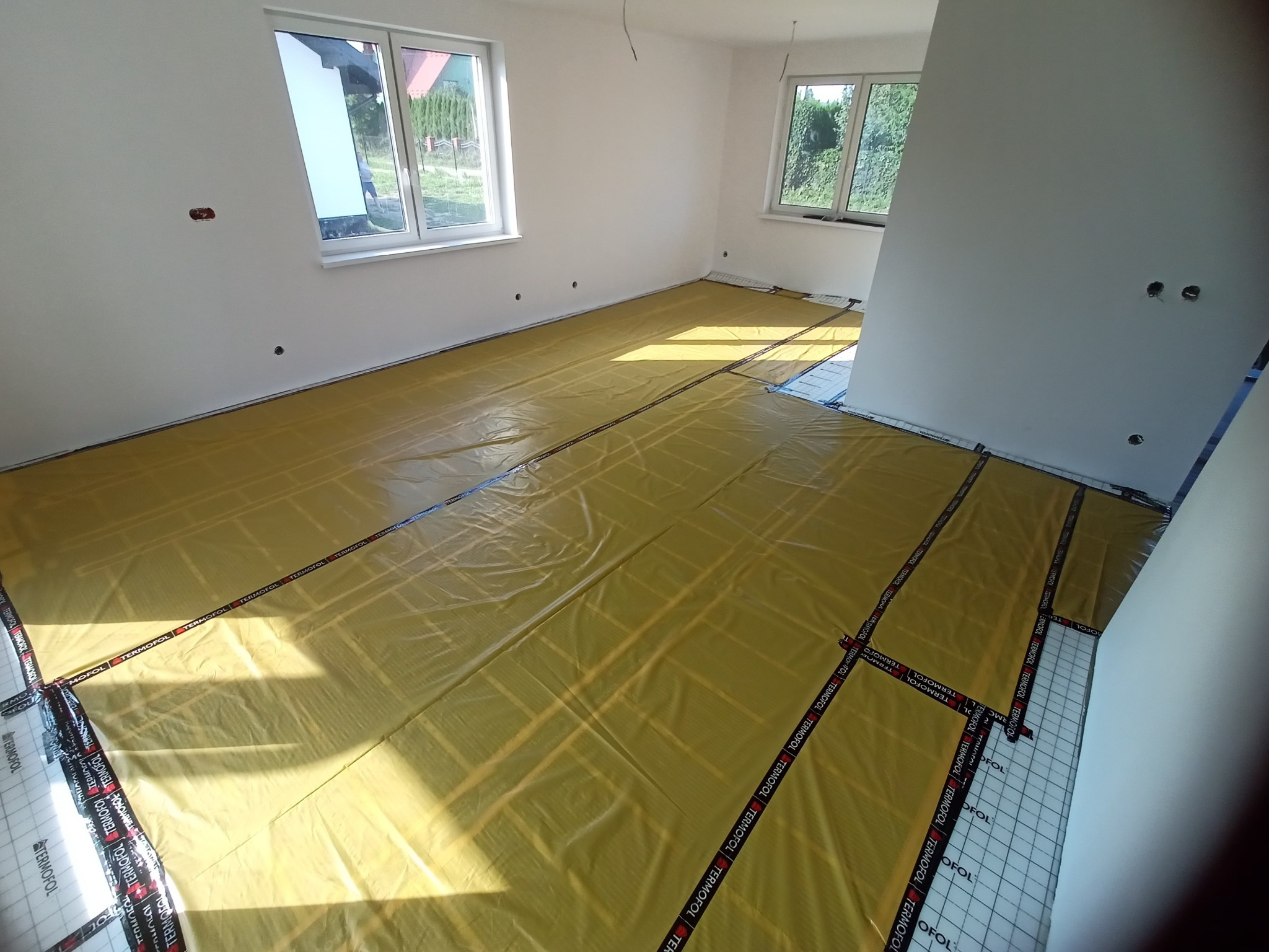 Żółta folia grzewcza rozłożona na podłodze w jasnym pokoju z dwoma oknami, przygotowana do dalszych prac wykończeniowych, widoczne taśmy montażowe z logo Termofol.