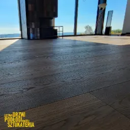 Ciemna, drewniana podłoga z widocznym rysunkiem słojów, oświetlona naturalnym światłem w nowoczesnym wnętrzu z dużym oknem i widokiem na zewnątrz.