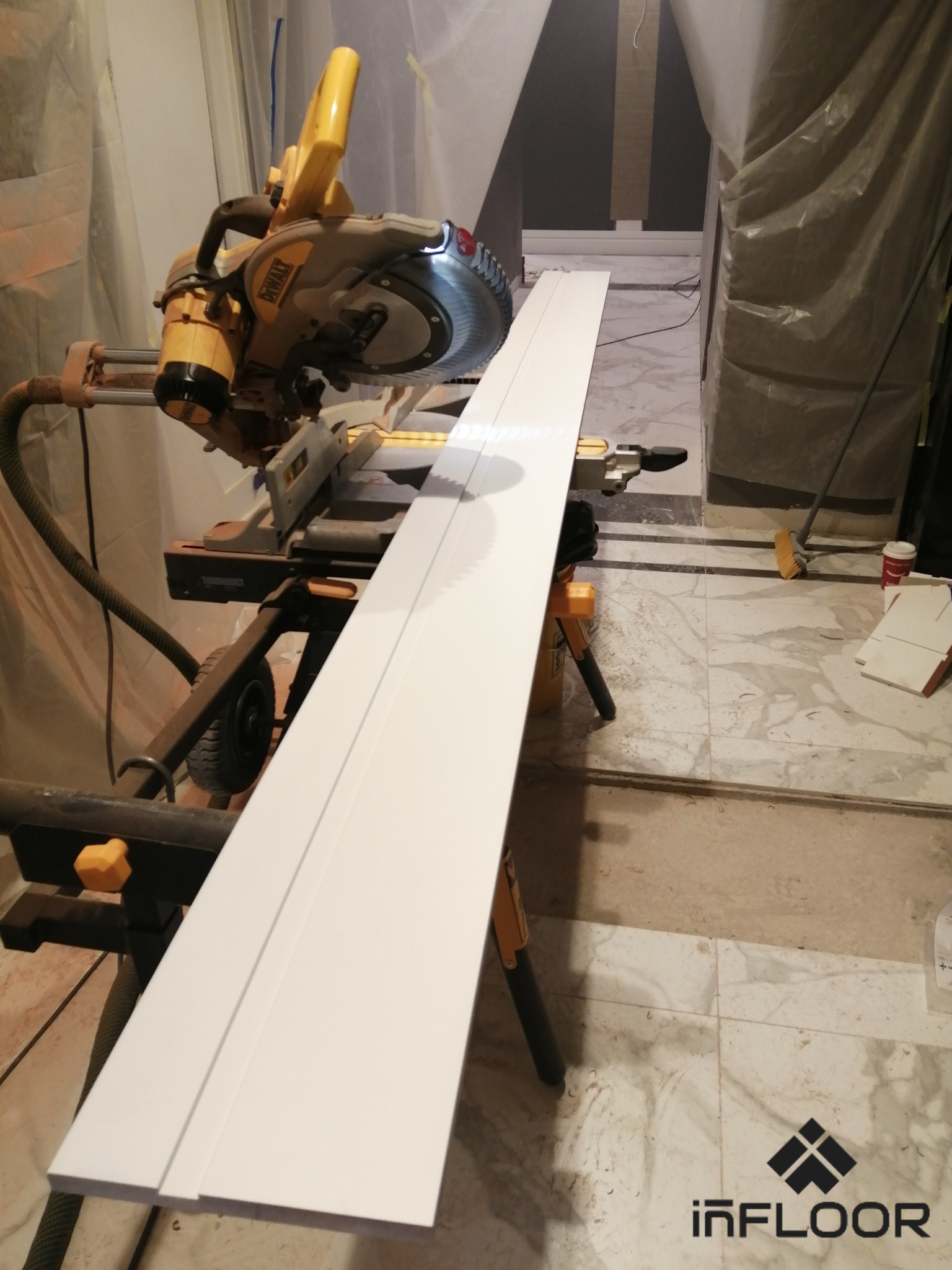 Biała listwa przypodłogowa przygotowana do cięcia na pile ukośnicy DeWalt, widoczne zabezpieczenia folią malarską.