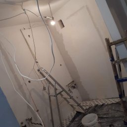 WOMEN'S TEAM PERFECT-BUD-CLEANING WIOLA&JOLA JOLANTA DOBROWOLSKA - Wnętrze w trakcie prac wykończeniowych: widoczne przewody elektryczne, drabina, częściowo wygładzona ściana z otworem, wiadro i worek z zaprawą na podłodze zabezpieczonej folią.