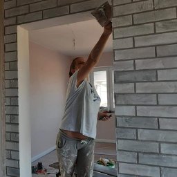 WOMEN'S TEAM PERFECT-BUD-CLEANING WIOLA&JOLA JOLANTA DOBROWOLSKA - Kobieta w ubrudzonych farbą spodniach i koszulce nakłada zaprawę na ścianę wykończoną szarą cegłą dekoracyjną przy użyciu kielni, widoczne rusztowanie i narzędzia w pomieszczeniu z oknem.