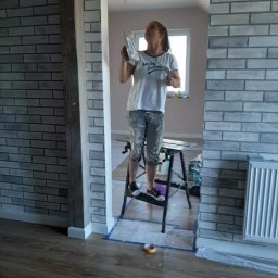 WOMEN'S TEAM PERFECT-BUD-CLEANING WIOLA&JOLA JOLANTA DOBROWOLSKA - Kobieta na drabince szpachluje ścianę imitującą cegłę w przejściu między pokojami, widoczne narzędzia i zabezpieczona podłoga.