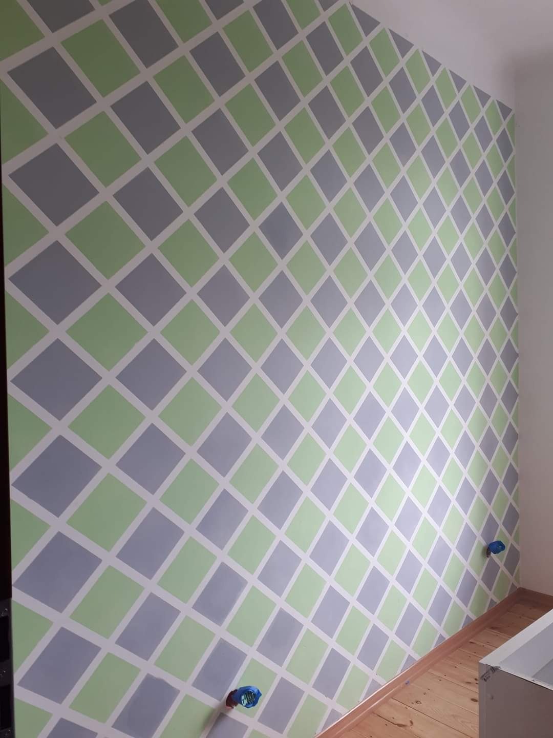 Ściana w pokoju pomalowana w szaro-zielone romby z białymi paskami, widoczne gniazdka elektryczne.