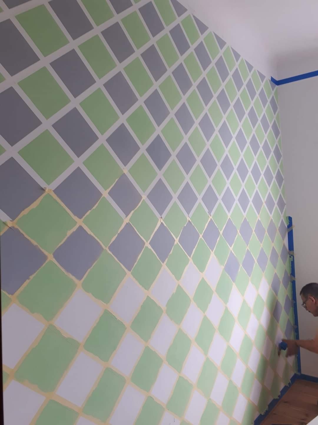 Malowanie ściany w romby z użyciem taśmy malarskiej, kolory: biały, szary i pistacjowy, widoczny fragment podłogi i malarz podczas pracy.