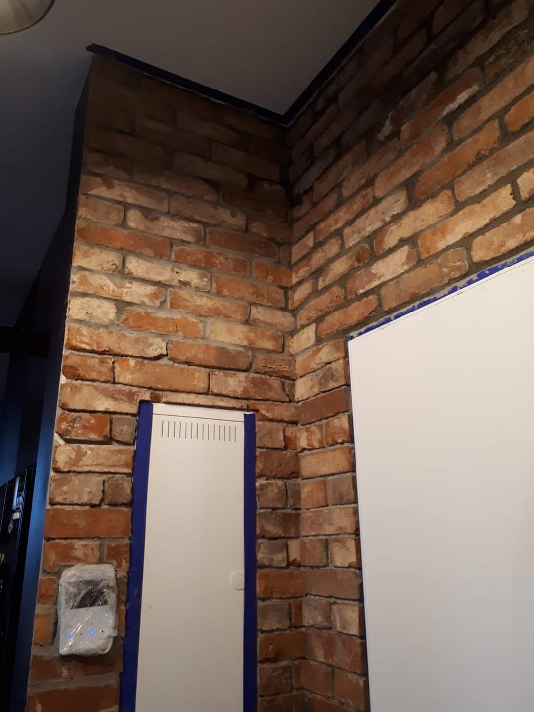 Ściana z cegły klinkierowej w rogu pomieszczenia, zabezpieczona niebieską taśmą malarską wokół białych drzwi rewizyjnych i skrzynki elektrycznej, widoczne niedoskonałości fug.