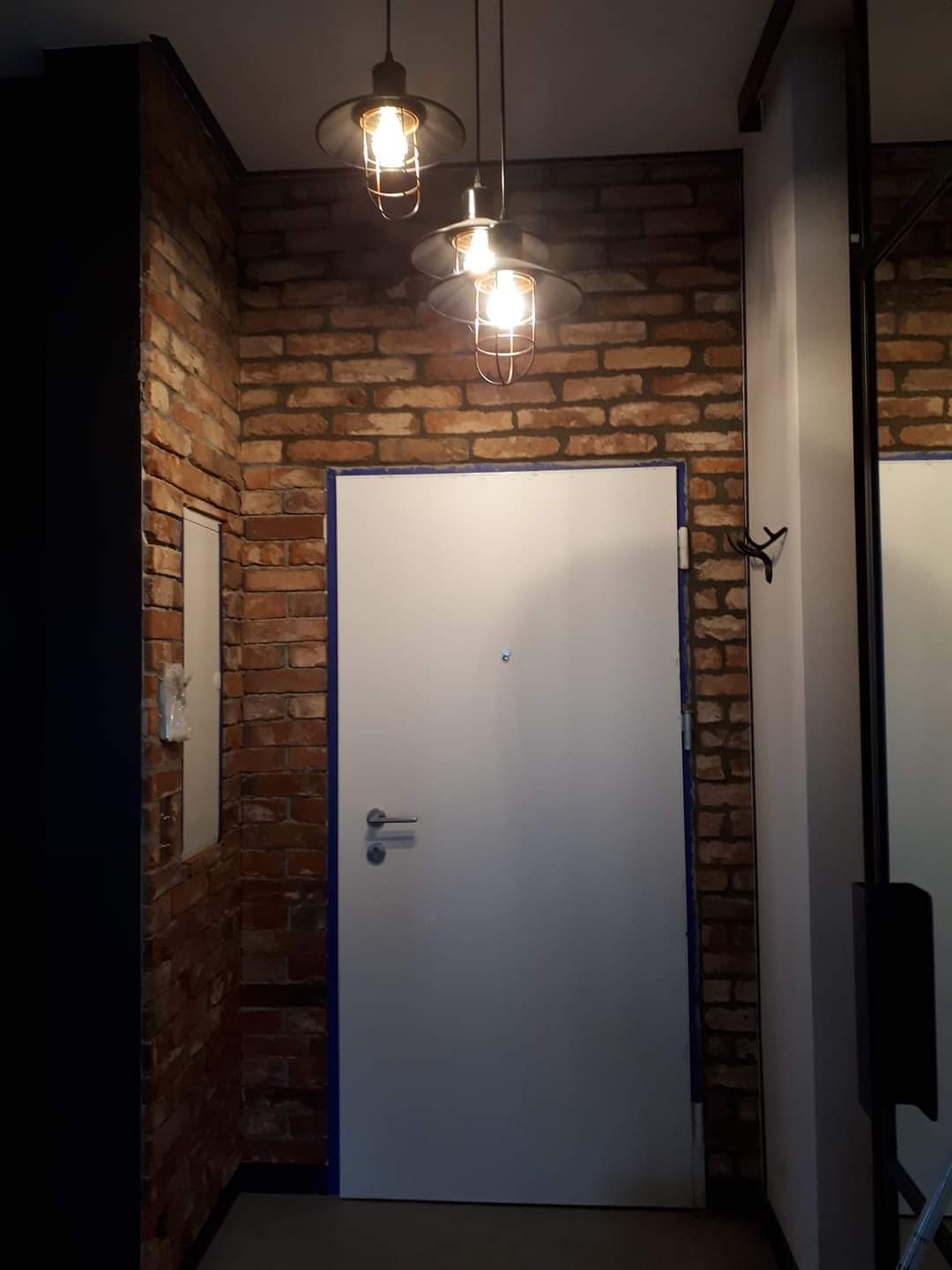 Białe drzwi wejściowe z niebieską ramą w murze z czerwonej cegły, oświetlone trzema wiszącymi lampami w stylu industrialnym.