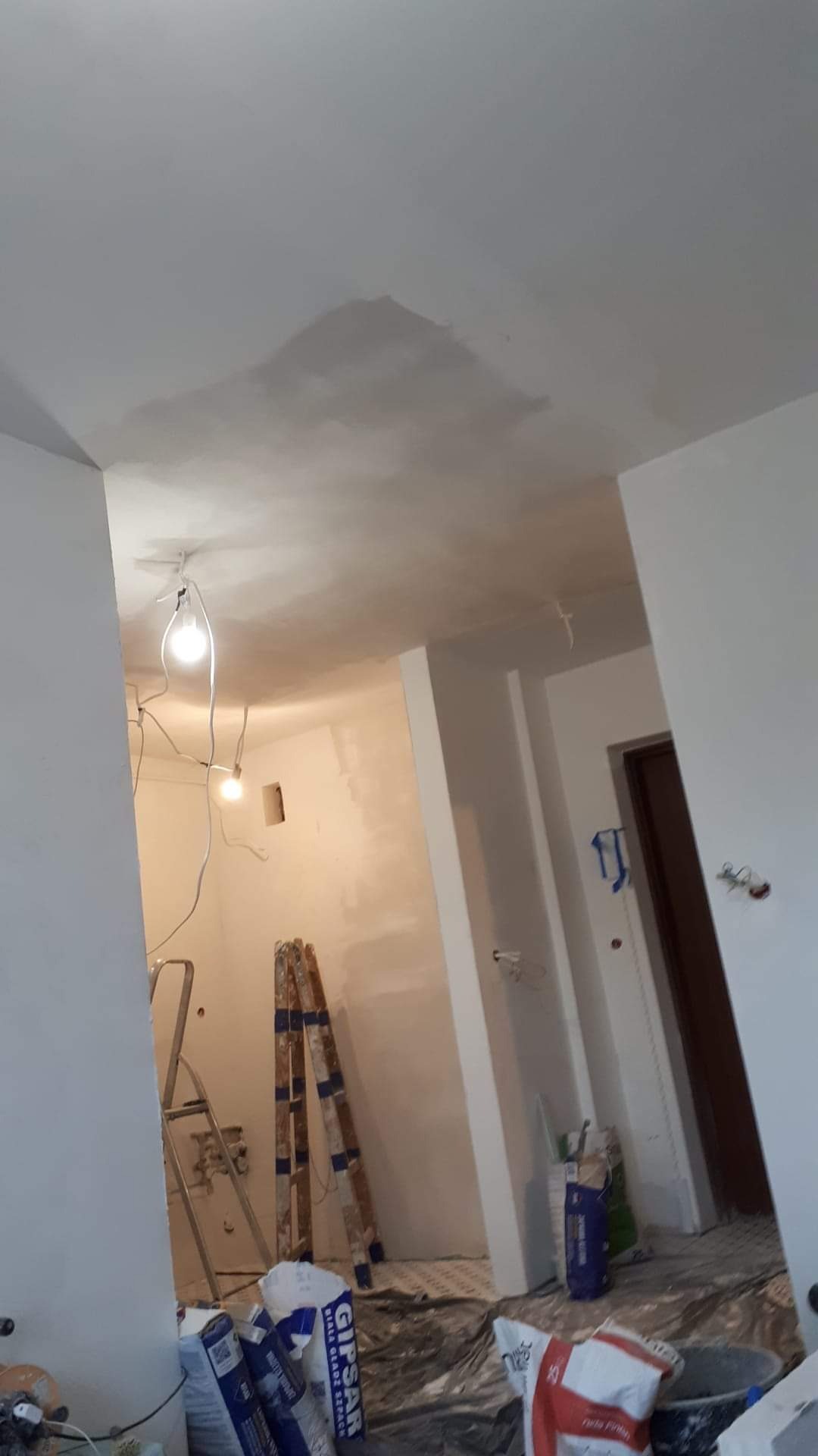 Wnętrze w trakcie remontu: widoczne niedoskonałości na suficie po położeniu gładzi, przewody z żarówkami zwisające z sufitu, drabiny opierające się o ścianę, worki z gładzią gipsową na podłodze...