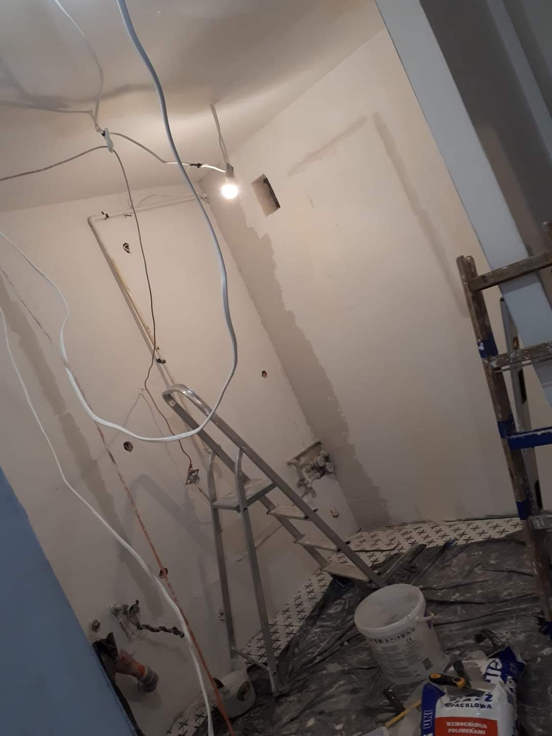 Wnętrze w trakcie prac wykończeniowych: widoczne przewody elektryczne, drabina, częściowo wygładzona ściana z otworem, wiadro i worek z zaprawą na podłodze zabezpieczonej folią.