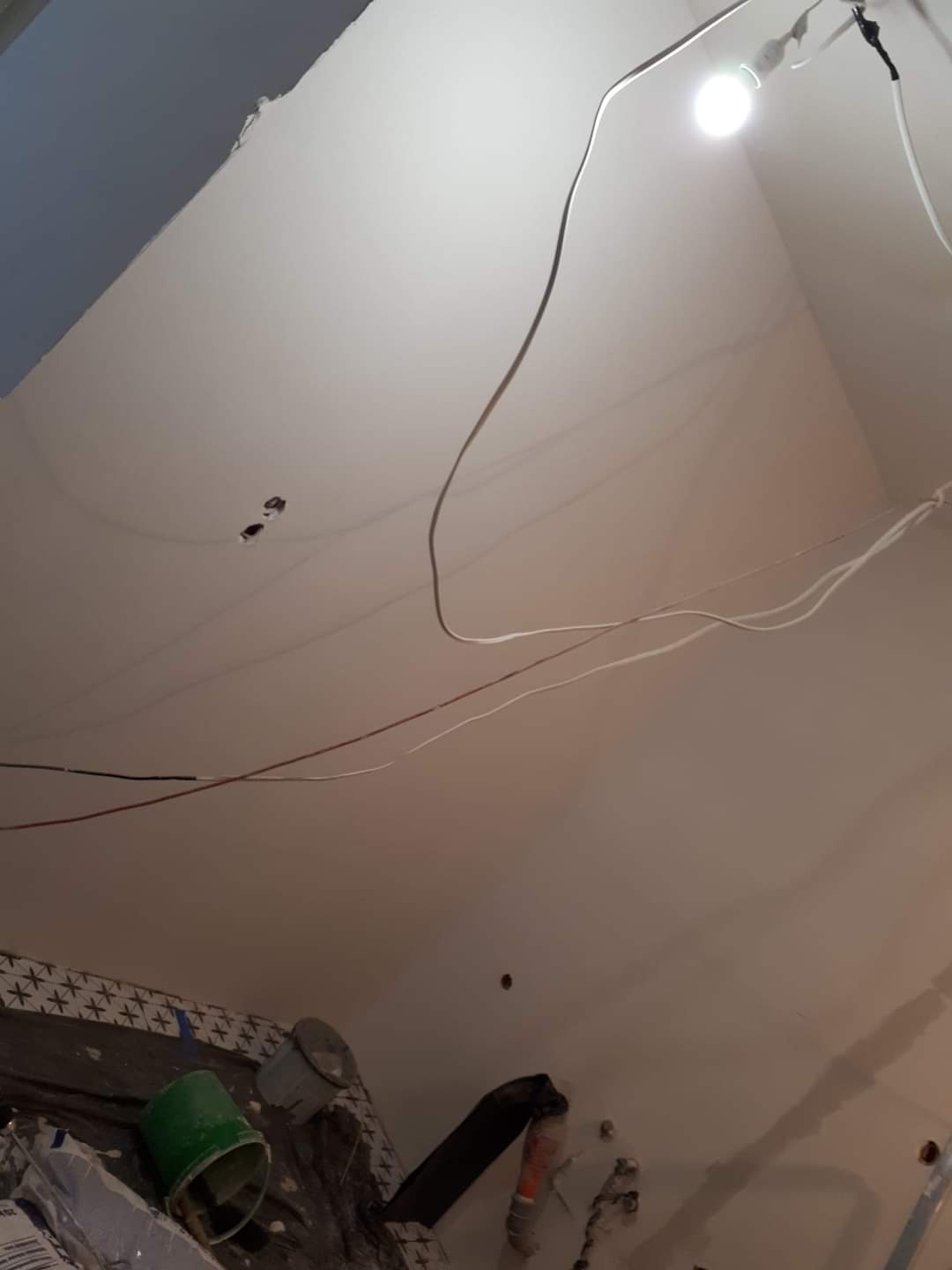 Widok w górę na sufit w trakcie prac wykończeniowych, widoczne wiszące kable, prowizoryczne oświetlenie żarówką oraz fragment ściany z narzędziami i materiałami budowlanymi w dolnej części kadru.