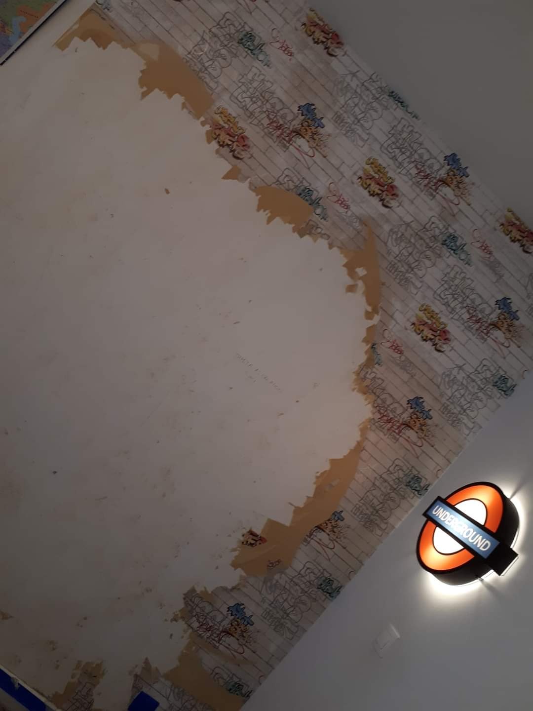 Fragment ściany w trakcie usuwania tapety z motywem graffiti i cegieł, obok lampa w stylu londyńskiego metra.