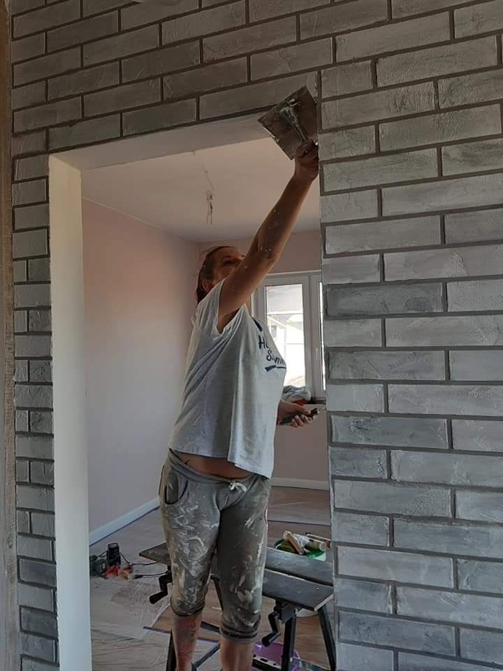 Kobieta w ubrudzonych farbą spodniach i koszulce nakłada zaprawę na ścianę wykończoną szarą cegłą dekoracyjną przy użyciu kielni, widoczne rusztowanie i narzędzia w pomieszczeniu z oknem.