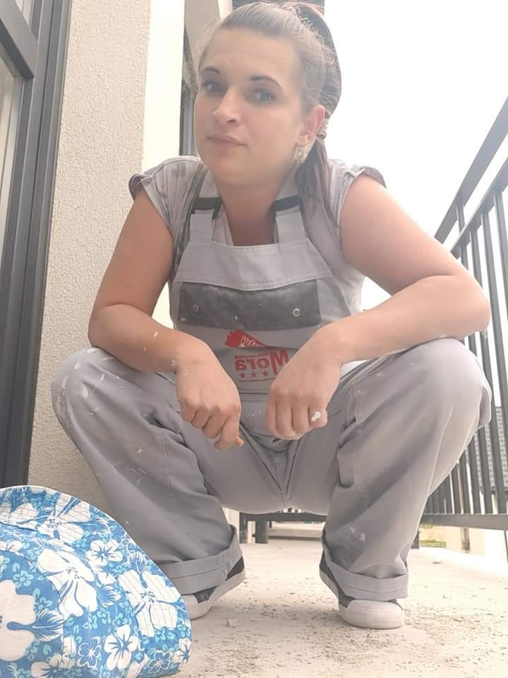 Kobieta w roboczym kombinezonie z plamami farby, kucająca na balkonie, z torbą z kwiatowym wzorem obok, prawdopodobnie po zakończeniu pracy malarskiej.