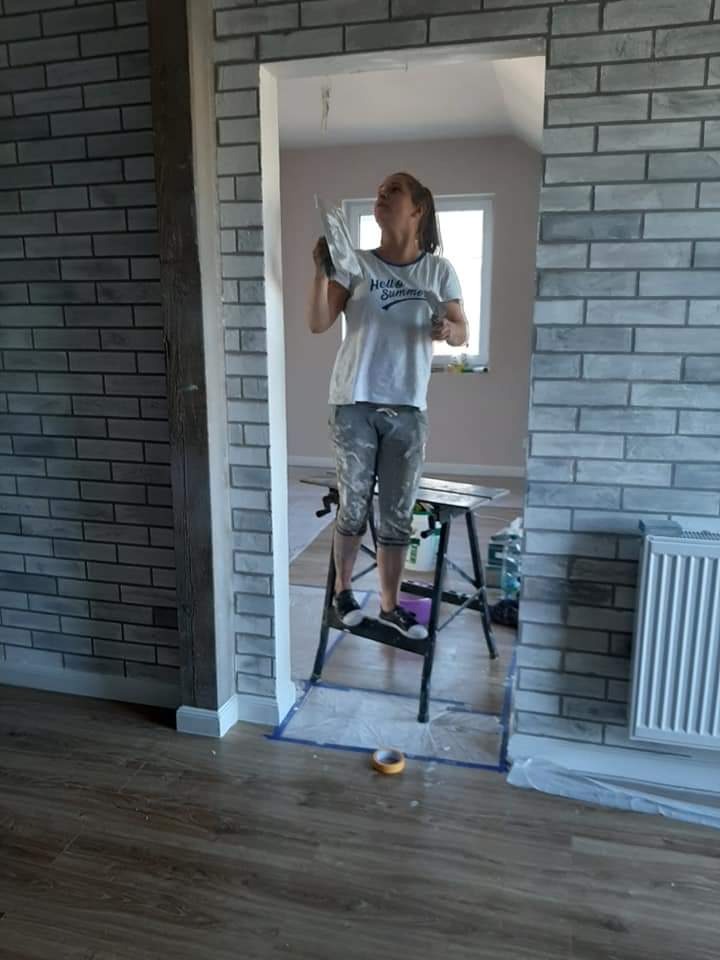 Kobieta na drabince szpachluje ścianę imitującą cegłę w przejściu między pokojami, widoczne narzędzia i zabezpieczona podłoga.