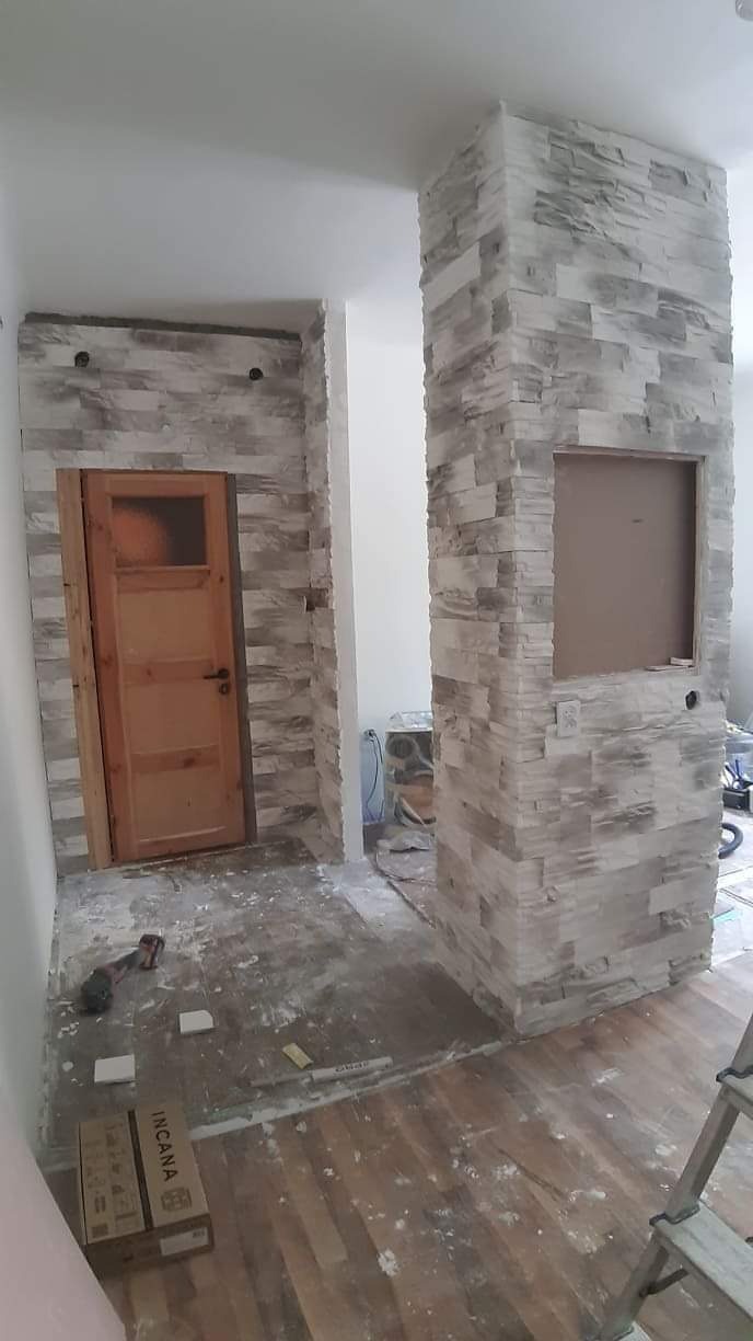 Ściana i filar obłożone dekoracyjnym kamieniem elewacyjnym w odcieniach szarości, widoczna surowa drewniana ościeżnica i fragment podłogi z resztkami zaprawy, karton po produkcie Incana oraz drabina.