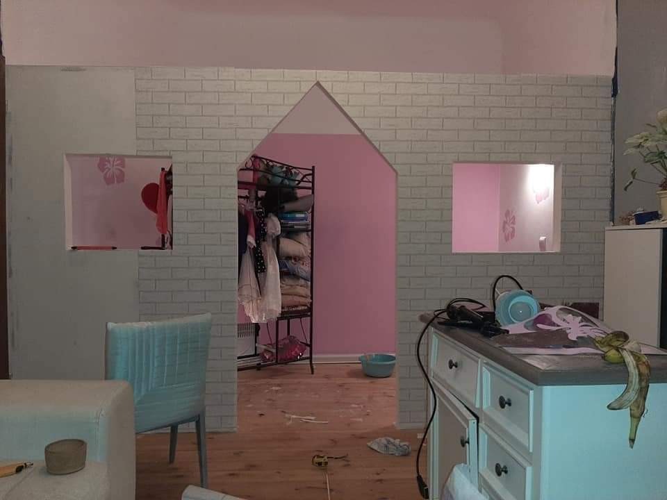 Wnętrze pokoju dziecięcego w trakcie tworzenia zabudowy przypominającej domek z szarej cegły, z dwoma okienkami i wejściem w kształcie domku, widoczny bałagan i narzędzia pracy.