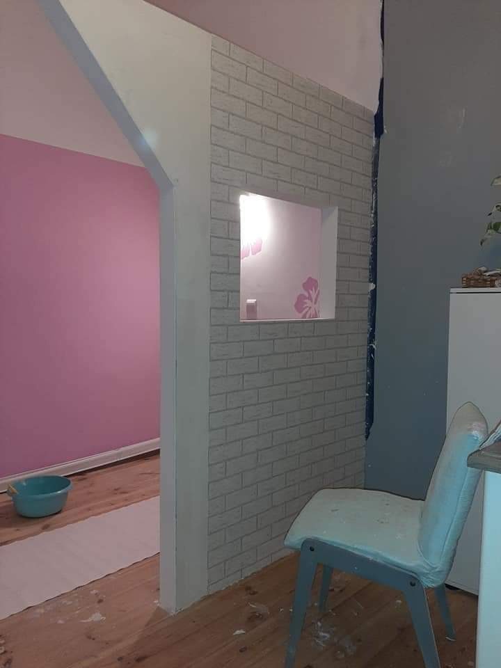 Fragment pokoju w trakcie prac wykończeniowych: ściana z imitacji cegły z wnęką, różowa ściana w tle, podłoga częściowo zabezpieczona folią, krzesło w rogu.