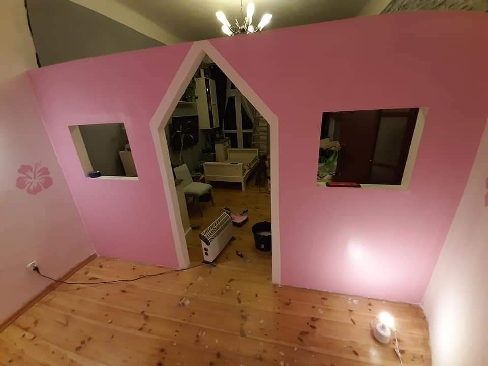 Pokój dziecięcy z różową ścianą w kształcie domku z oknami i wejściem, widoczny grzejnik elektryczny i lampka na podłodze z desek.