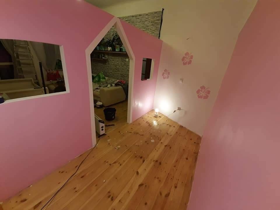 Pokój dziecięcy w trakcie aranżacji: różowe ściany, drewniana podłoga, dekoracyjne kwiatowe wzory na ścianie, widoczne elementy umeblowania i akcesoria w tle, częściowo ukończona konstrukcja...