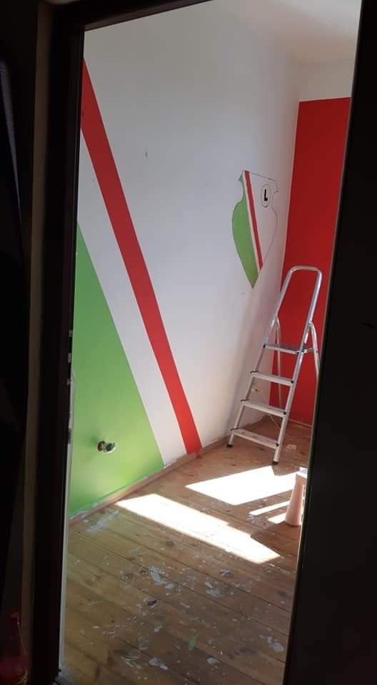 Pokój po malowaniu z motywem klubowym: biała ściana z ukośnymi pasami w kolorach zielonym i czerwonym, częściowo widoczne logo klubu, aluminiowa drabina oparta o ścianę, drewniana podłoga z plamami...