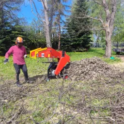 Pracownik w kasku i okularach ochronnych obsługuje czerwoną rębarkę do gałęzi, obok sterta zrębków, w tle przyczepka i trawnik z żółtymi kwiatami.