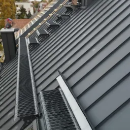 Montaż nowoczesnego, grafitowego dachu z blachy na rąbek stojący z systemem wentylacji kalenicowej. Widoczny dekarz pracujący przy kominie.